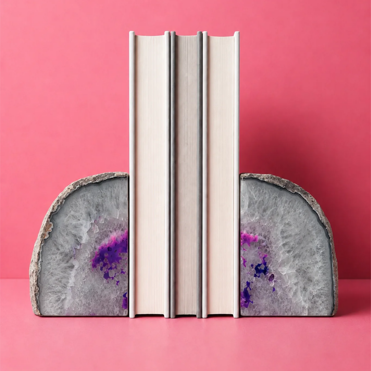 Two agate geode bookends with purple and gray patterns hold up two neutral-colored books against a solid pink background. 