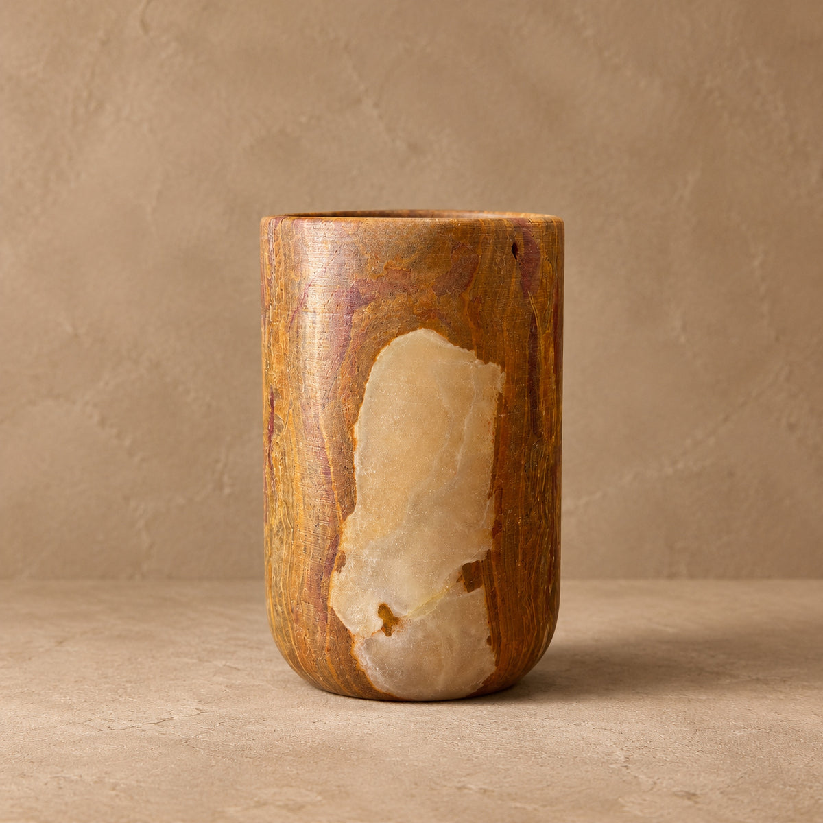 Saffron onyx vessel with rust-red banding and a large translucent honey window, a straight cylinder on a rounded foot, against a warm plaster backdrop