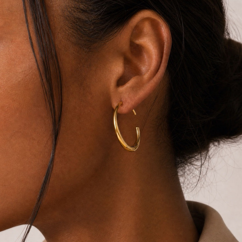 A close-up side profile of a person wearing a polished gold hoop earring, showing the ear, hair, and neck against a plain light background.
