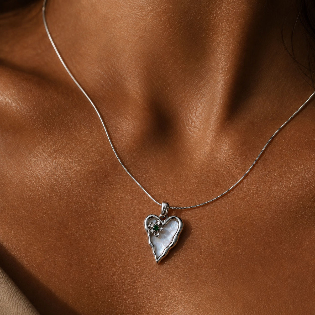 A close-up of a delicate silver chain necklace worn on a model's neck, featuring a heart-shaped pendant with a white mother-of-pearl center and a small clear gemstone.

Silver molten-edge heart pendant with white shell pearl and green cubic zirconia flower, worn on a fine snake chain against sunlit skin at the collarbone
