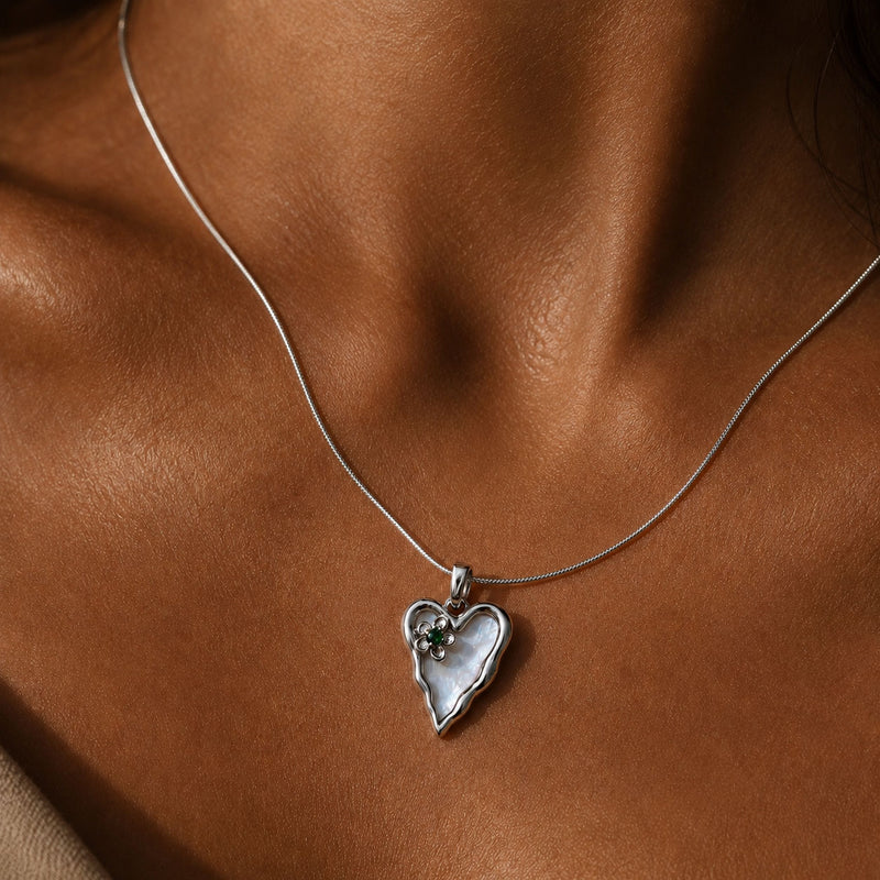 A close-up of a delicate silver chain necklace worn on a model's neck, featuring a heart-shaped pendant with a white mother-of-pearl center and a small clear gemstone.

Silver molten-edge heart pendant with white shell pearl and green cubic zirconia flower, worn on a fine snake chain against sunlit skin at the collarbone
