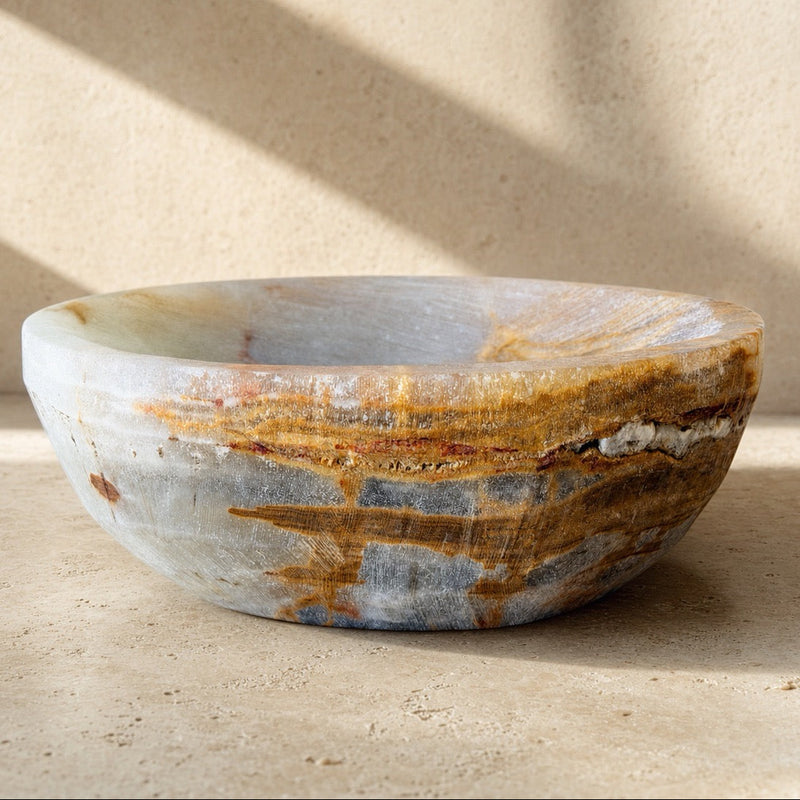 A round stone vessel bowl with natural marbled patterns in gray, beige, and brown, set against a textured beige wall with soft diagonal shadows.

Side profile of a slate grey onyx catchall bowl with an amber and rust vein sweeping across the surface, set against a sunlit stone wall.