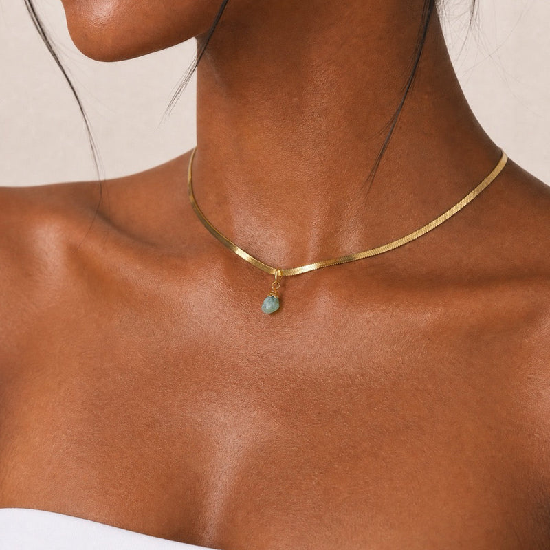 Close-up of a woman wearing a gold snake chain necklace choker 14 inch with a blue stone aquamarine pendant on a beige background