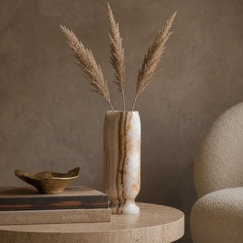 Tall hand-carved ivory onyx origin vase with vertical amber and rust banding, holding pampas grass on a travertine side table beside stacked books, a brass dish and a boucle chair