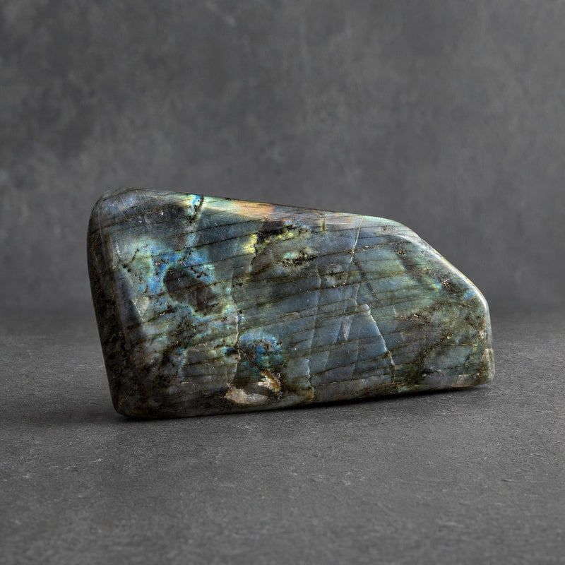 Teal and gold labradorite freeform, polished, at a three-quarter angle: blue and teal flash with a gold band across the face, on charcoal.