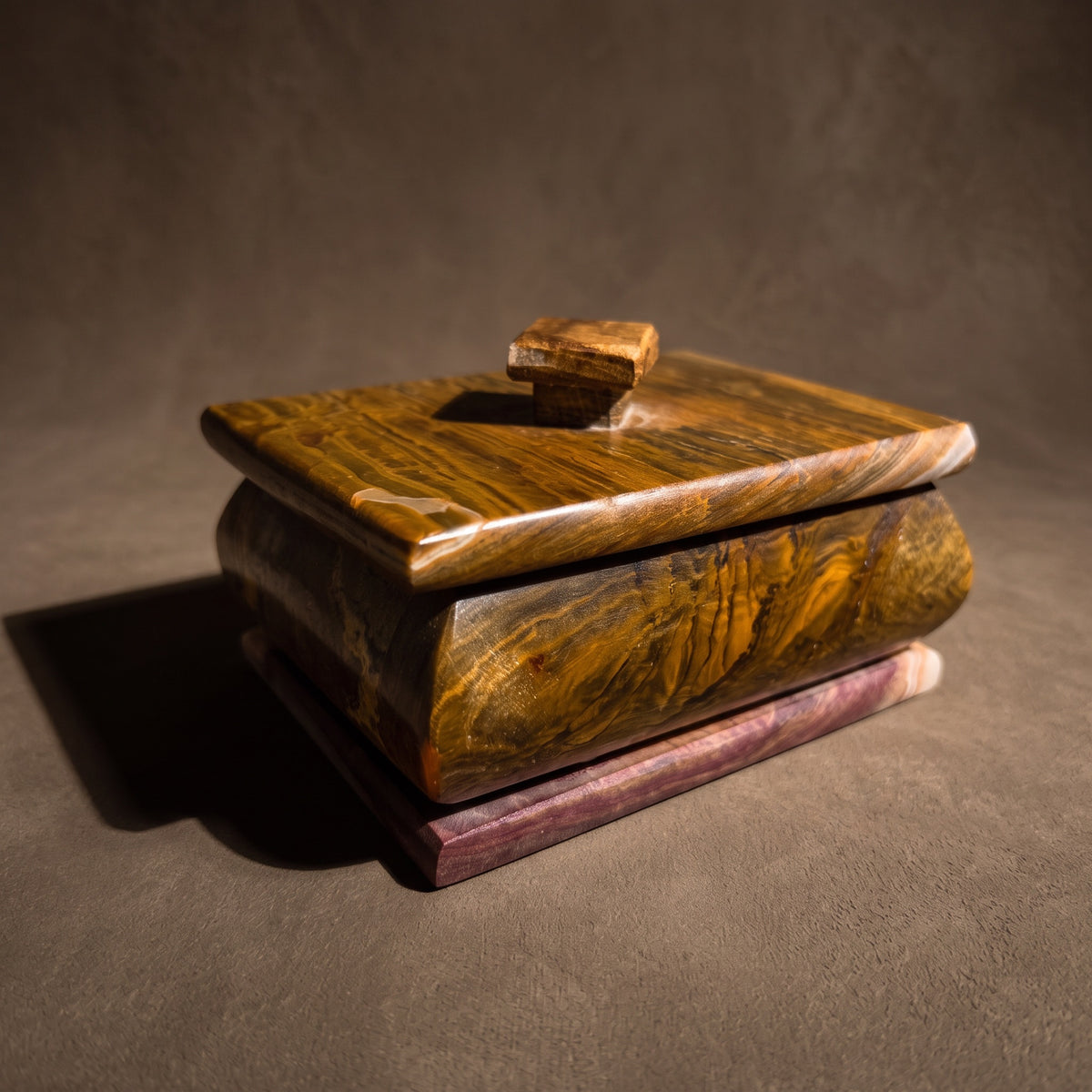 Tobacco, caramel and wine onyx keepsake box with lid at a three-quarter angle: wood-grain banding across the lid and bulged body, a wine-red stepped base and a small square stone knob, in raking light