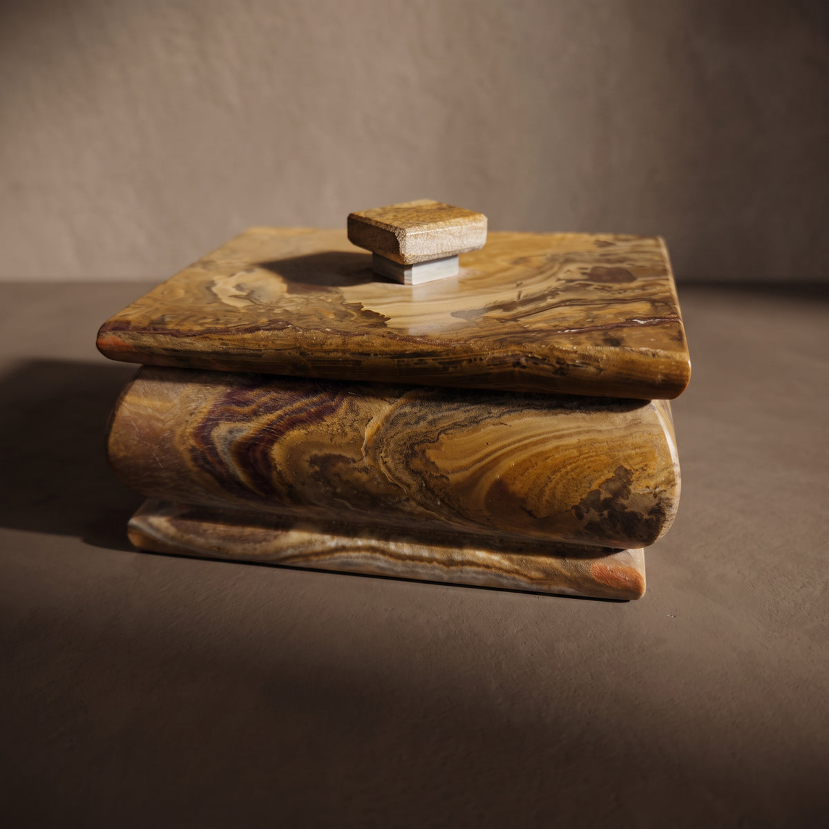 Side profile of the hand-carved onyx keepsake box, toffee and cream swirls with a mulberry streak across the rounded body, under an overhanging lid with a square knob