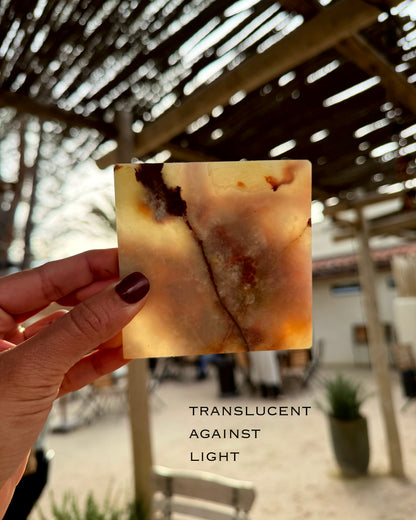 Hand holding a translucent square onyx marble stone coaster with a blurred outdoor background