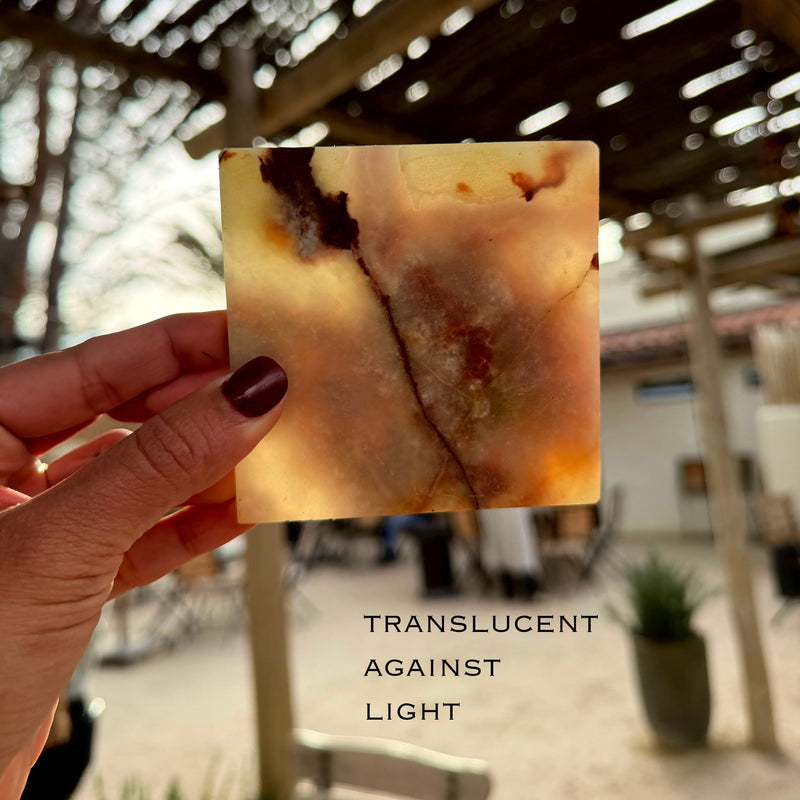 Hand holding a translucent square onyx marble stone coaster with a blurred outdoor background