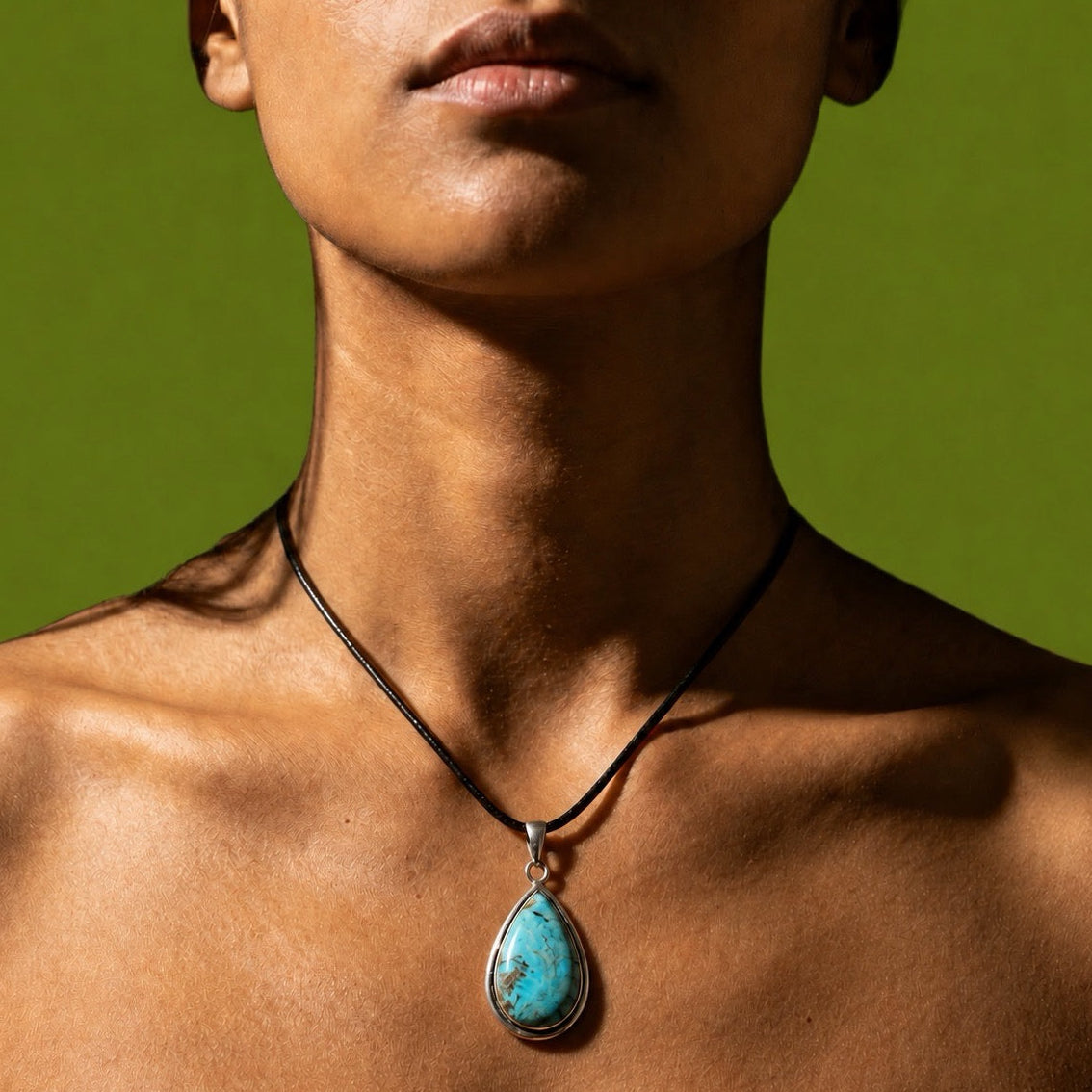 A close-up of a person's neck and upper chest wearing a black cord necklace with a teardrop-shaped turquoise pendant in a silver bezel setting, against a solid olive green background.