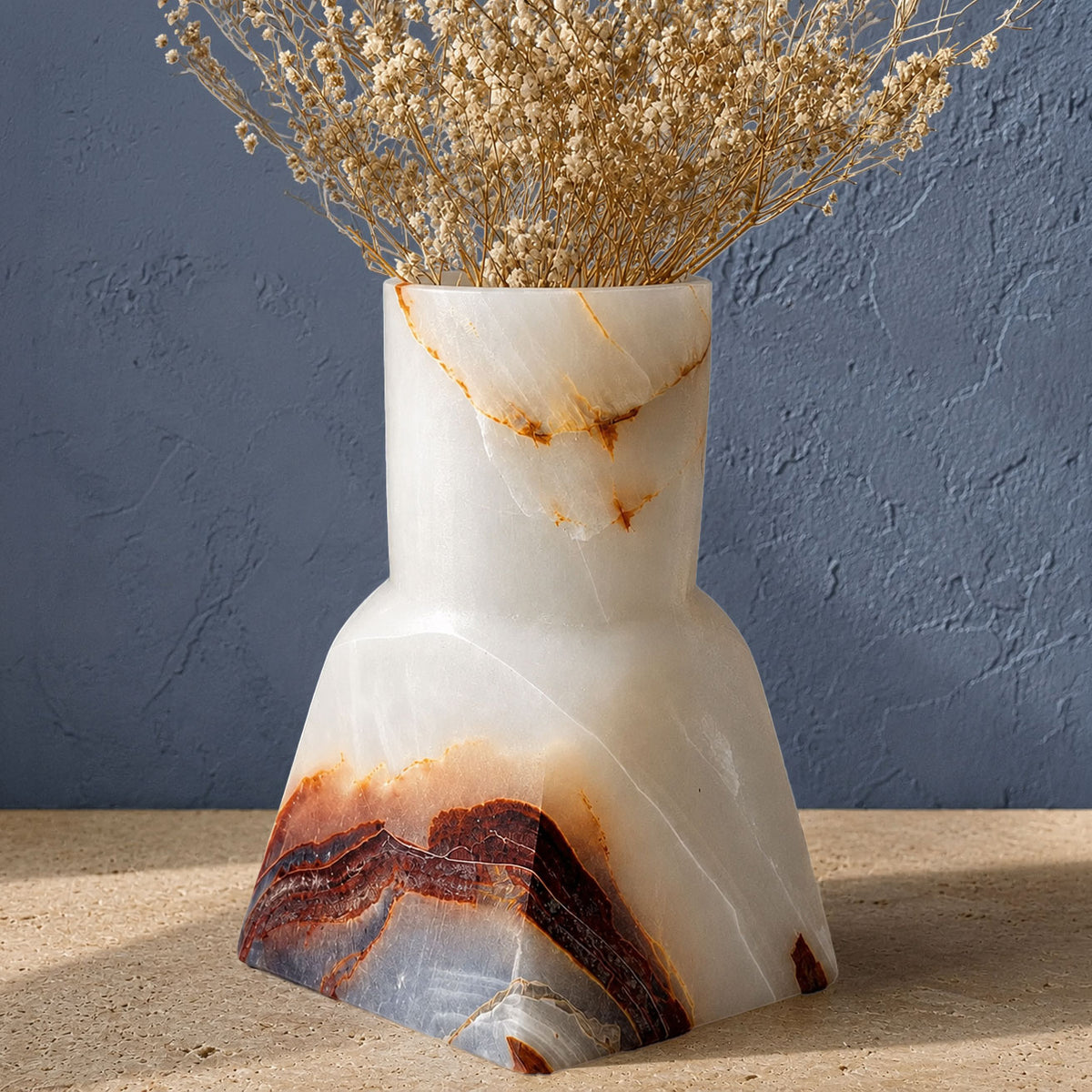 A sculptural white onyx vase with natural brown and orange veining, holding dried beige flowers, set on a light stone surface against a textured blue-gray wall.