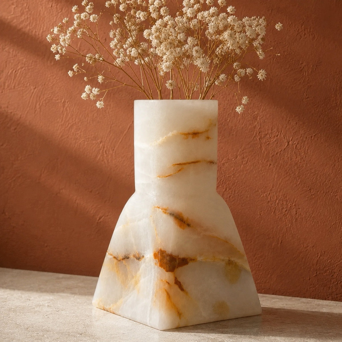 A white onyx marble vase with natural brown and gold veining, holding dried white baby's breath flowers, set on a light stone surface against a textured terracotta wall.