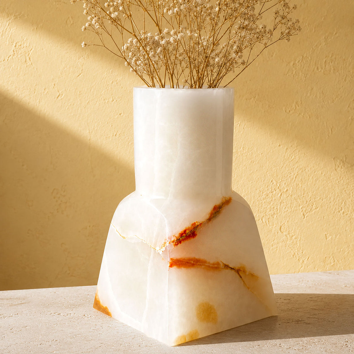 A white onyx vase with natural orange-brown veining, holding dried baby's breath stems, set on a light stone surface against a textured beige wall with warm sunlight and shadows.