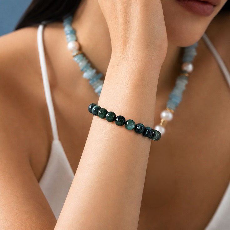 A close-up of a woman's wrist wearing a beaded bracelet made of dark green and blue-green round beads, with a matching light blue and white beaded necklace also visible.