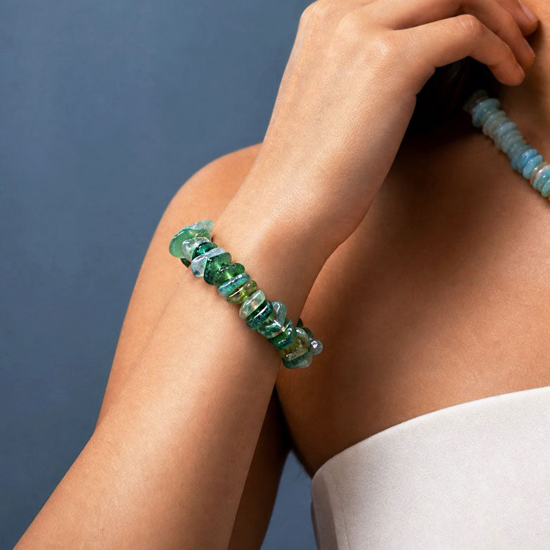 A close-up lifestyle shot of a person wearing a chunky bracelet made of polished green and blue-green gemstone beads, with a matching necklace partially visible.