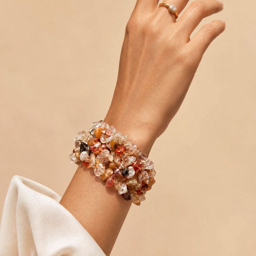 A close-up of a person's wrist wearing a chunky, multi-strand crystal bracelet with clear, orange, brown, and black stones, styled with a white sleeve and a gold ring on the finger, set against a solid beige background.