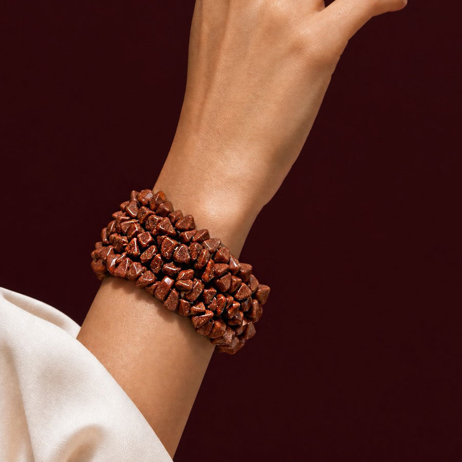 A close-up of a person's wrist wearing a chunky multi-strand bracelet made of polished reddish-brown stone chips, set against a dark burgundy background.