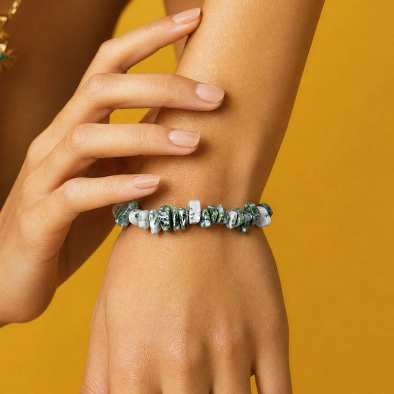 A close-up of a person's wrist wearing a beaded stretch bracelet made of irregular green and white marbled stones, set against a solid mustard yellow background.